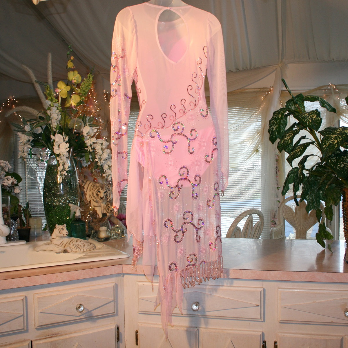 back view of stunning pink Latin-Rhythm dress. Its delicate soft pink stretch mesh is adorned with a mesmerizing scroll design, highlighted by two shades of glimmering pink rhinestones in a beautiful scroll design on the back, upper bodice, and left sheer side. The extensive hand beading in the skirting adds the perfect touch of elegance