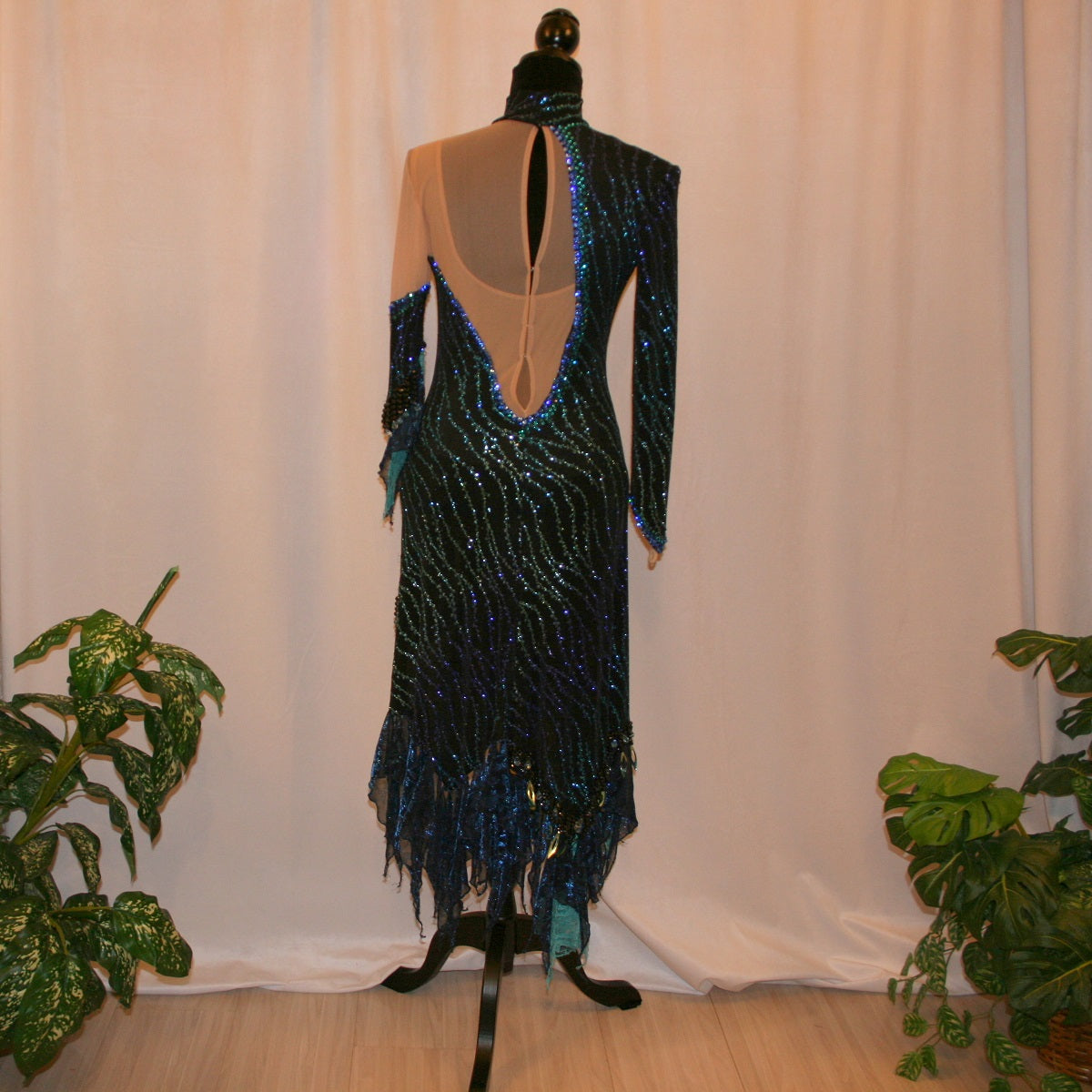 back view of black Latin-rhythm dress sparkles plenty on the dance floor with a stunning black glitter fabric featuring a wavy turquoise, aqua & royal blue design, complete with black hand-beading, aqua teardrop shape beads, spangles, and intricate flounces in various fabrics of deep shimmering blue, turquoise, aqua & black.