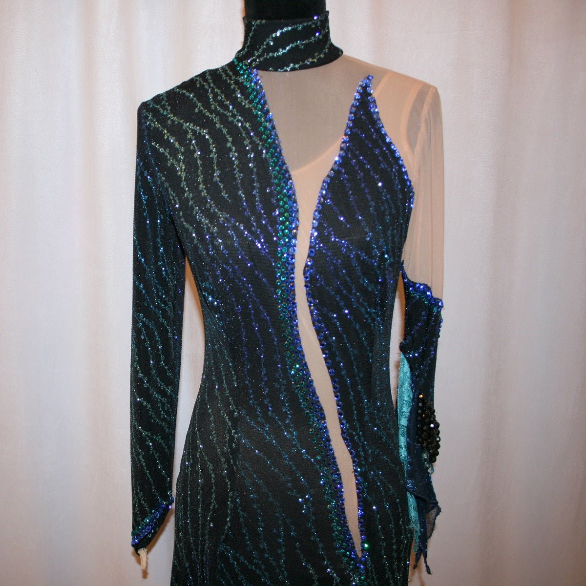 close up front view of black Latin-rhythm dress sparkles plenty on the dance floor with a stunning black glitter fabric featuring a wavy turquoise, aqua & royal blue design, complete with black hand-beading, aqua teardrop shape beads, spangles, and intricate flounces in various fabrics of deep shimmering blue, turquoise, aqua & black.