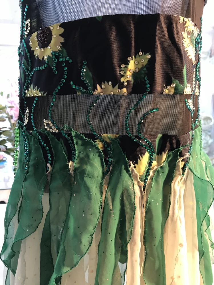 Close up view of details on all around ballroom dress that features yards of gorgeous soft yellow champagne sequin chiffon hand cut in large petal shapes for the skirting that is short in front & cascades longer in the back. Overlays of emerald flocked green chiffon skillfully hand crafted creating thin leaf shapes. Flows so beautifully for your foxtrots & waltzes! Lots of sparkle comes from the detailed rhinestone work