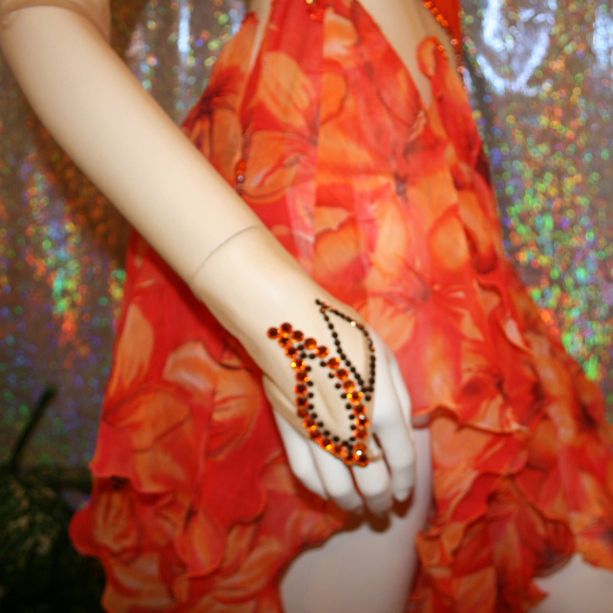 wrist detail view of Orange Latin-rhythm dress created on a nude illusion base of luxurious orange solid slinky with orange tropical print chiffon skirting is embellished with orange & bronze Swarovski rhinestone work & hand beading.