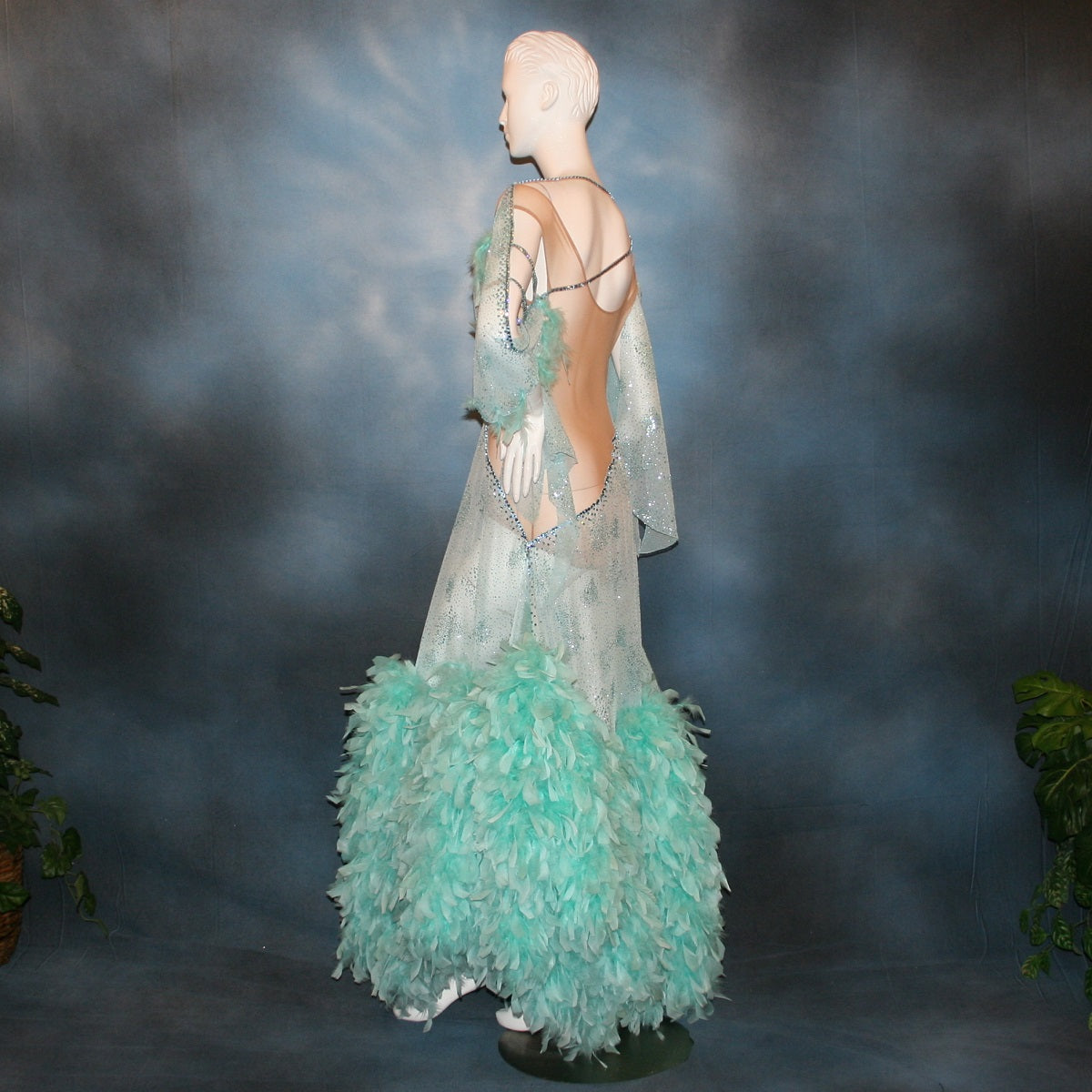 Crystal's Creations side view of Aqua blue ballroom dress created in gorgeous aqua blue glitter sheer mesh overlaid on a nude illusion base has lots of gorgeous aqua chandelle feathers, embellished with aquamarine Swarovski rhinestone work. This ballroom dress also features delicate Swarovski embellished strap detailing.