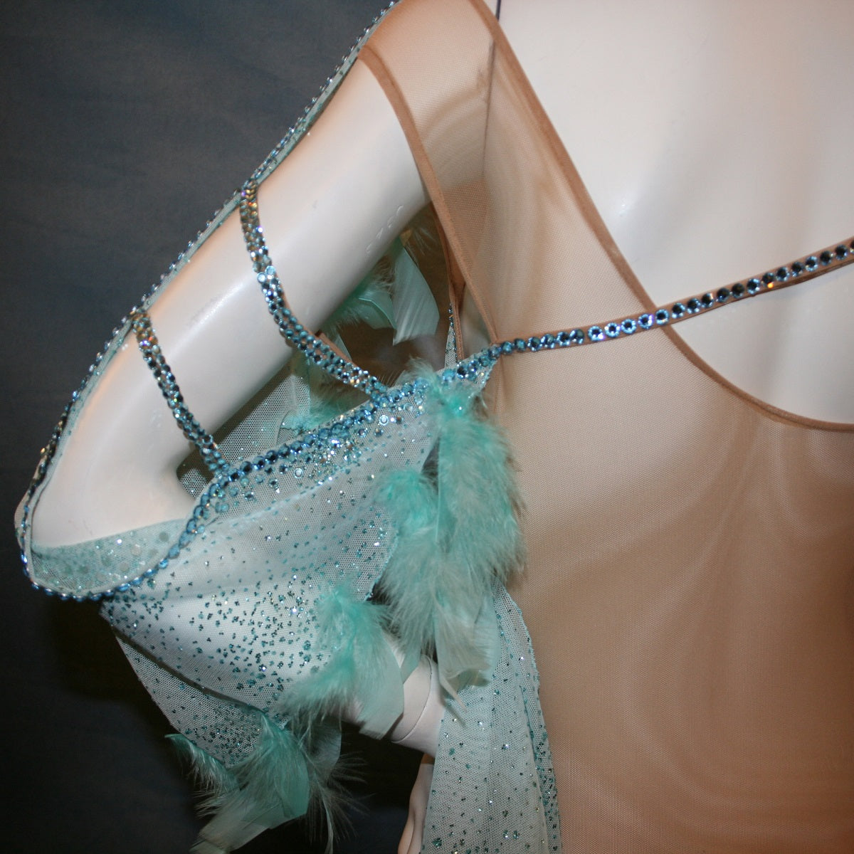 Crystal's Creations back shoulder view of Aqua blue ballroom dress created in gorgeous aqua blue glitter sheer mesh overlaid on a nude illusion base has lots of gorgeous aqua chandelle feathers, embellished with aquamarine Swarovski rhinestone work. This ballroom dress also features delicate Swarovski embellished strap detailing.