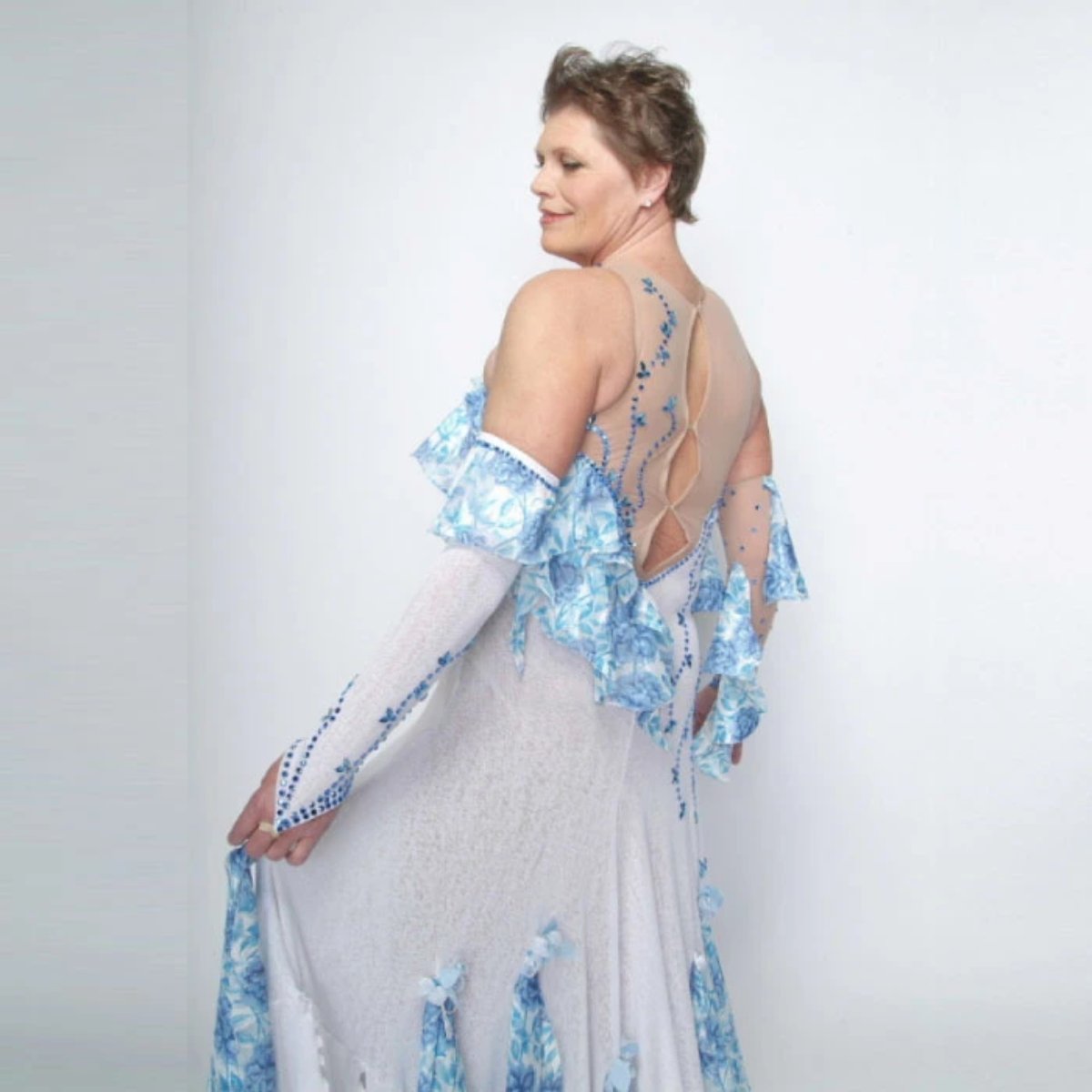 left upper side view of White ballroom dress with blue accents was created on nude illusion of a soft white knit with flounce accents of sky blue & white floral print satin, embellished with sapphire Swarovski stonework & miniature white silk roses.