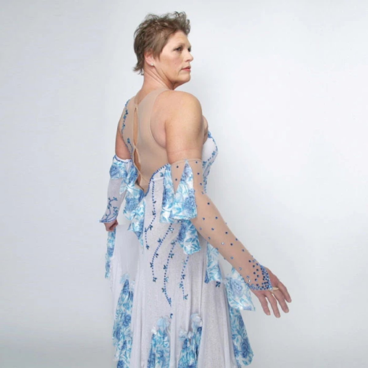 close upper side view of White ballroom dress with blue accents was created on nude illusion of a soft white knit with flounce accents of sky blue & white floral print satin, embellished with sapphire Swarovski stonework & miniature white silk roses.