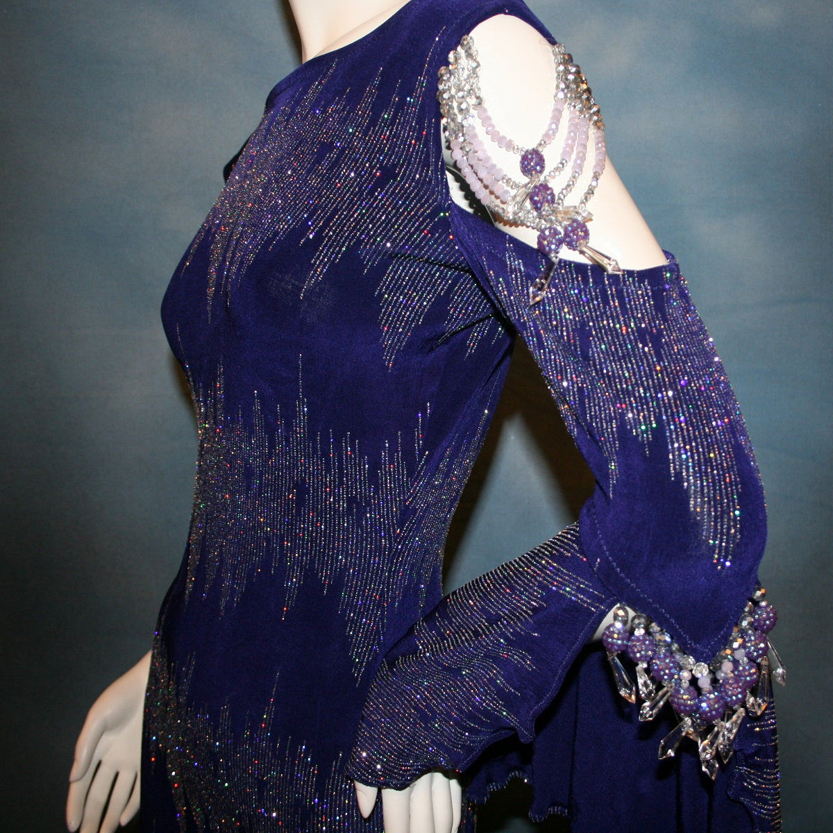 close view of details on Deep royal purple Latin/rhythm/tango dress created in deep royal purple glitter slinky with an awesome electrifying glitter pattern features one longe sleeve, with flair at the bottom, & another very interesting cold shoulder detailed one with hand beading & a flaired flounce.