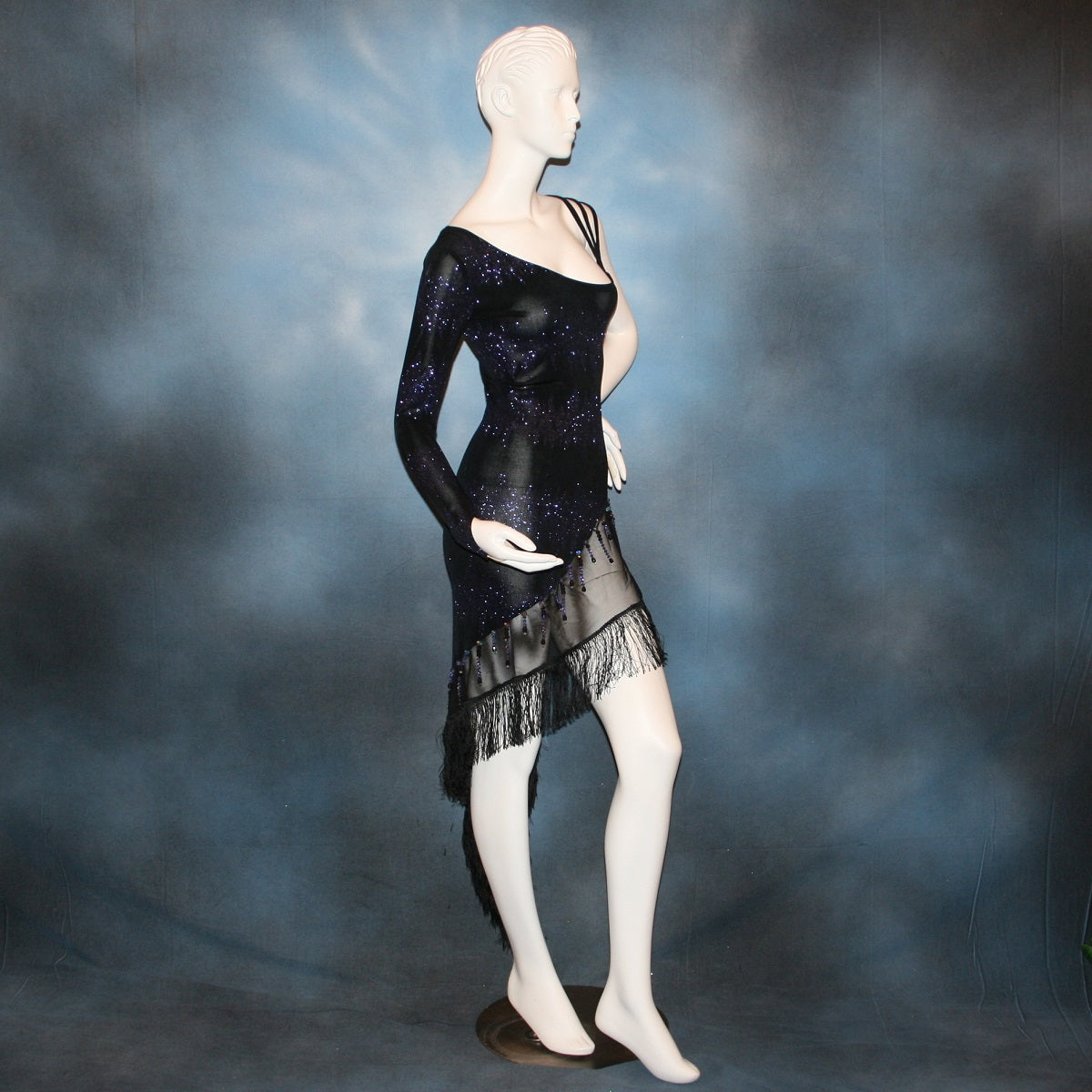side view of Latin/rhythm/tango dress created in black glitter slinky with an awesome electrifying tanzinite/perwinkle glitter pattern! It features one long sleeve, lattice work detailing up the left side, a short skirt line in front that angles down to a long peak in the back, with a sheer mesh inset, fringe & Swarovski hand beading in tanzanite & black.