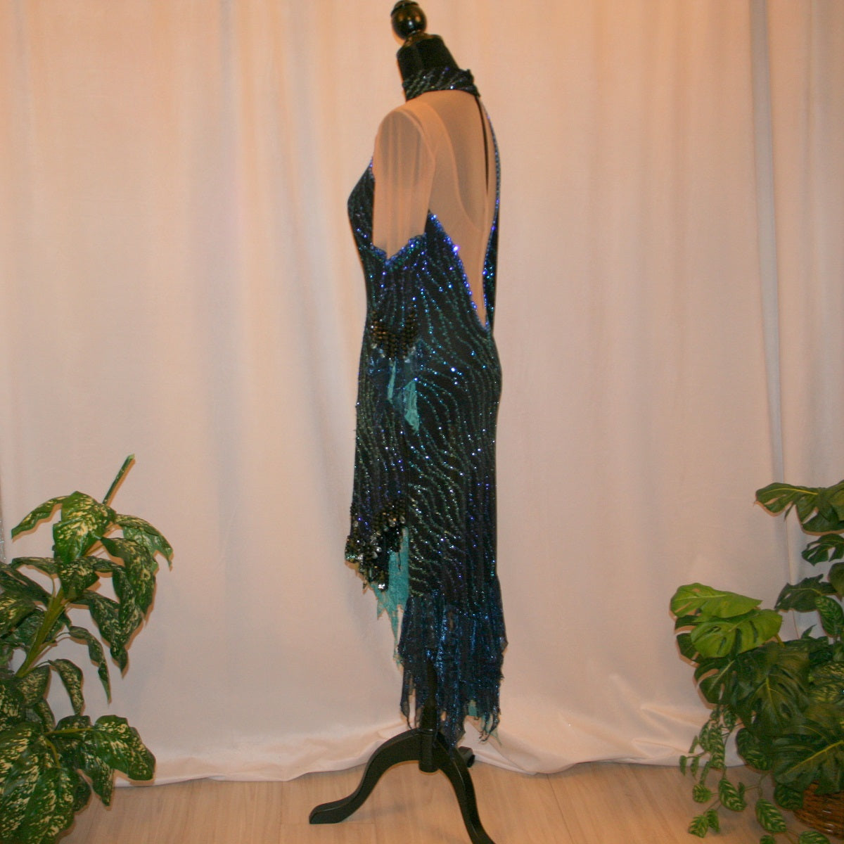 left side view of black Latin-rhythm dress sparkles plenty on the dance floor with a stunning black glitter fabric featuring a wavy turquoise, aqua & royal blue design, complete with black hand-beading, aqua teardrop shape beads, spangles, and intricate flounces in various fabrics of deep shimmering blue, turquoise, aqua & black. 