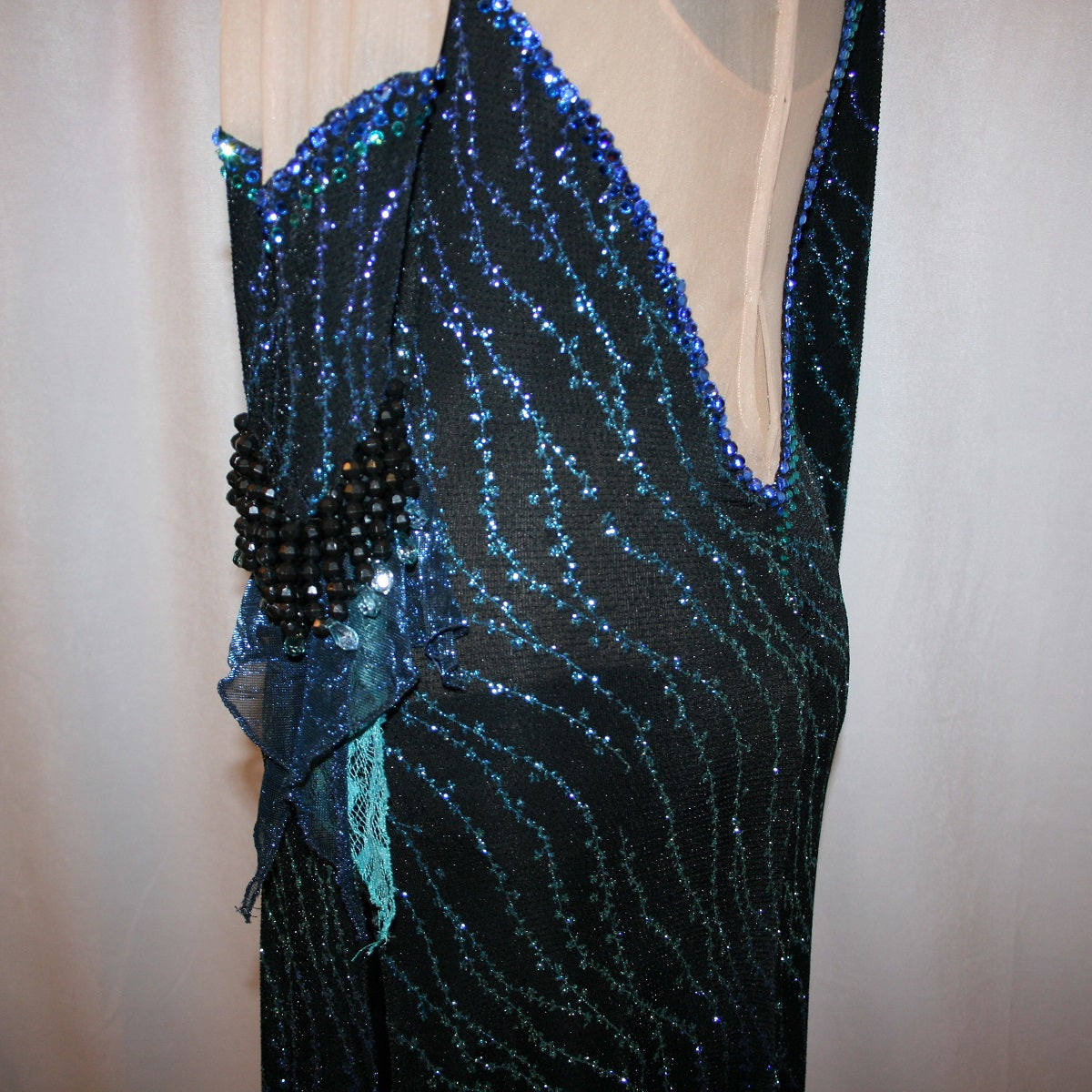 upper left side view of black Latin-rhythm dress sparkles plenty on the dance floor with a stunning black glitter fabric featuring a wavy turquoise, aqua & royal blue design, complete with black hand-beading, aqua teardrop shape beads, spangles, and intricate flounces in various fabrics of deep shimmering blue, turquoise, aqua & black. 