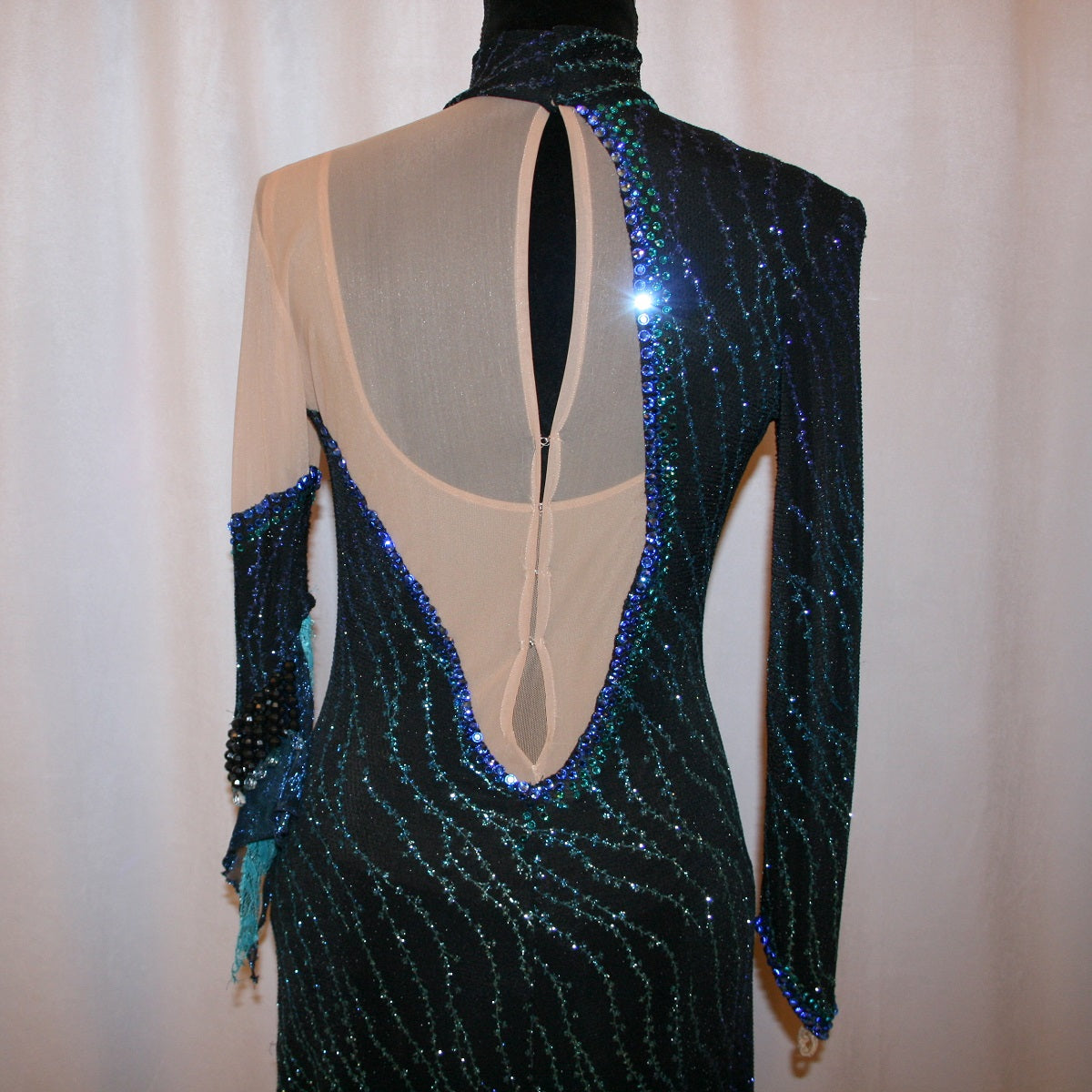 upper back view of black Latin-rhythm dress sparkles plenty on the dance floor with a stunning black glitter fabric featuring a wavy turquoise, aqua & royal blue design, complete with black hand-beading, aqua teardrop shape beads, spangles, and intricate flounces in various fabrics of deep shimmering blue, turquoise, aqua & black. 