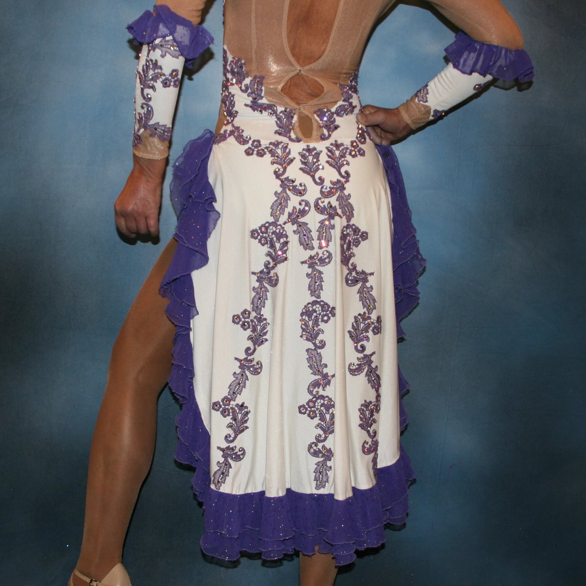 lower back view of White Latin/rhythm dress created with purple lace motifs embellished with crystal Aurora borealis Swarovski rhinestones overlaid on white lycra, and on nude illusion. The finishing touch are ruffles of glitter flecked chiffon.