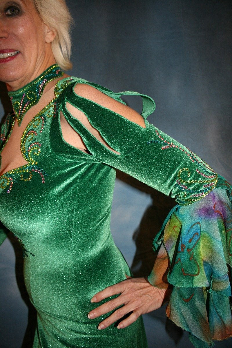 upper side view of Green Latin/rhythm dress was created in luxurious deep emerald green glitter stretch velvet with accent flounces of a green print chiffon. It features nude illusion cutout through the bodice, & is embellished with detailed Swarovski rhinestone work of peridot, emerald & orchid.