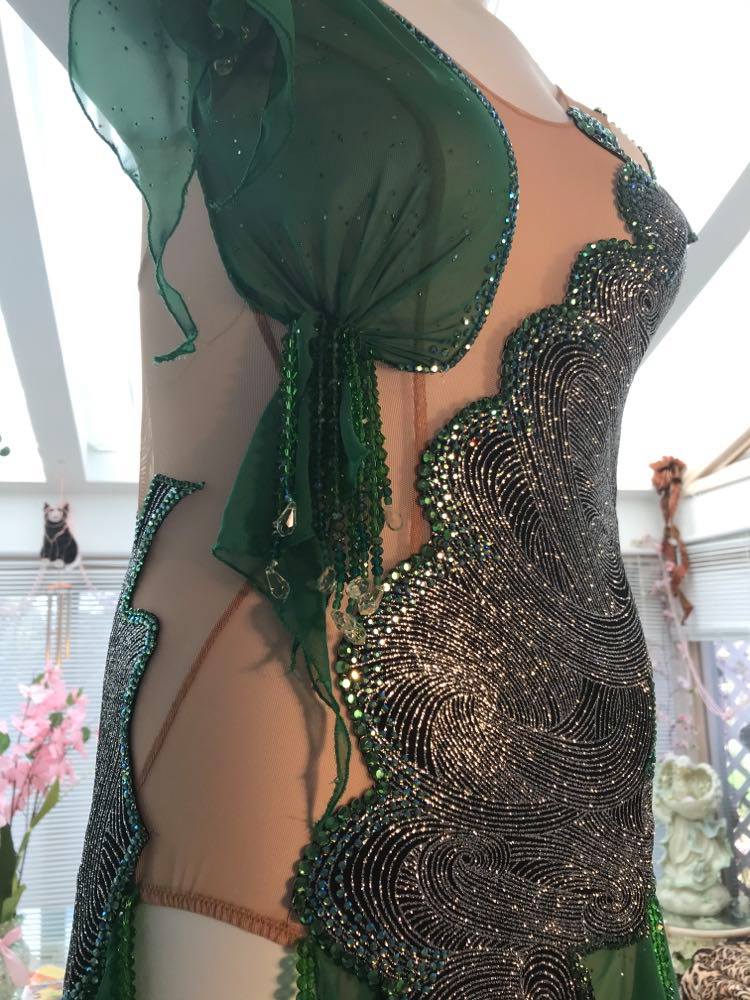 close up view of Latin/rhythm dress created of intricately cut & artistically placed silver swirls glitter slinky on a nude illusion base, with skirting & accent pieces of an emerald green