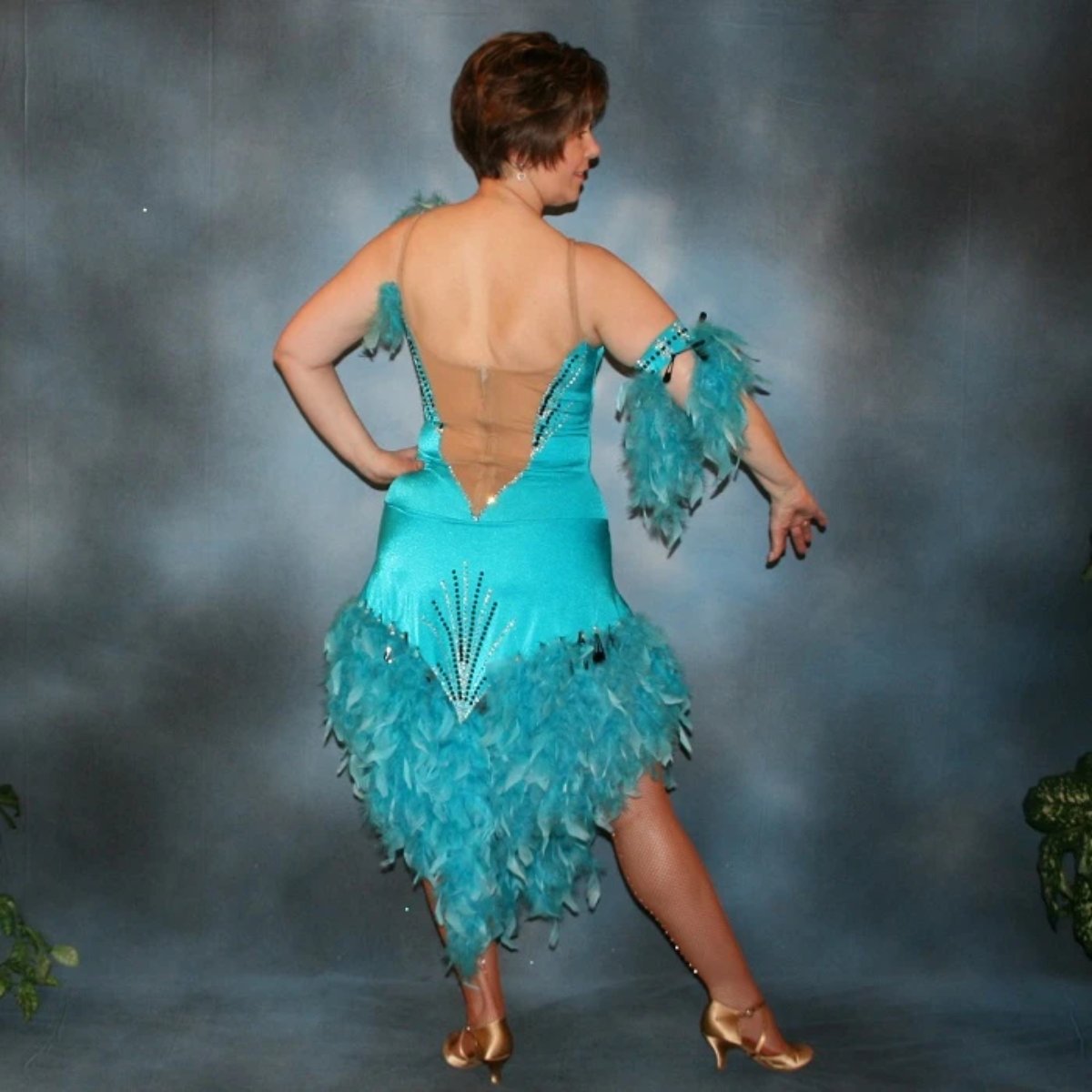 back view of Turquoise Latin/rhythm dance dress created in turquoise lycra on nude illusion, is embellished with crystal and jet black Swarovski rhinestones, with chandelle feathers and black spangles. The v styled skirt slits up high on both sides. Matching arm bands complete the look.