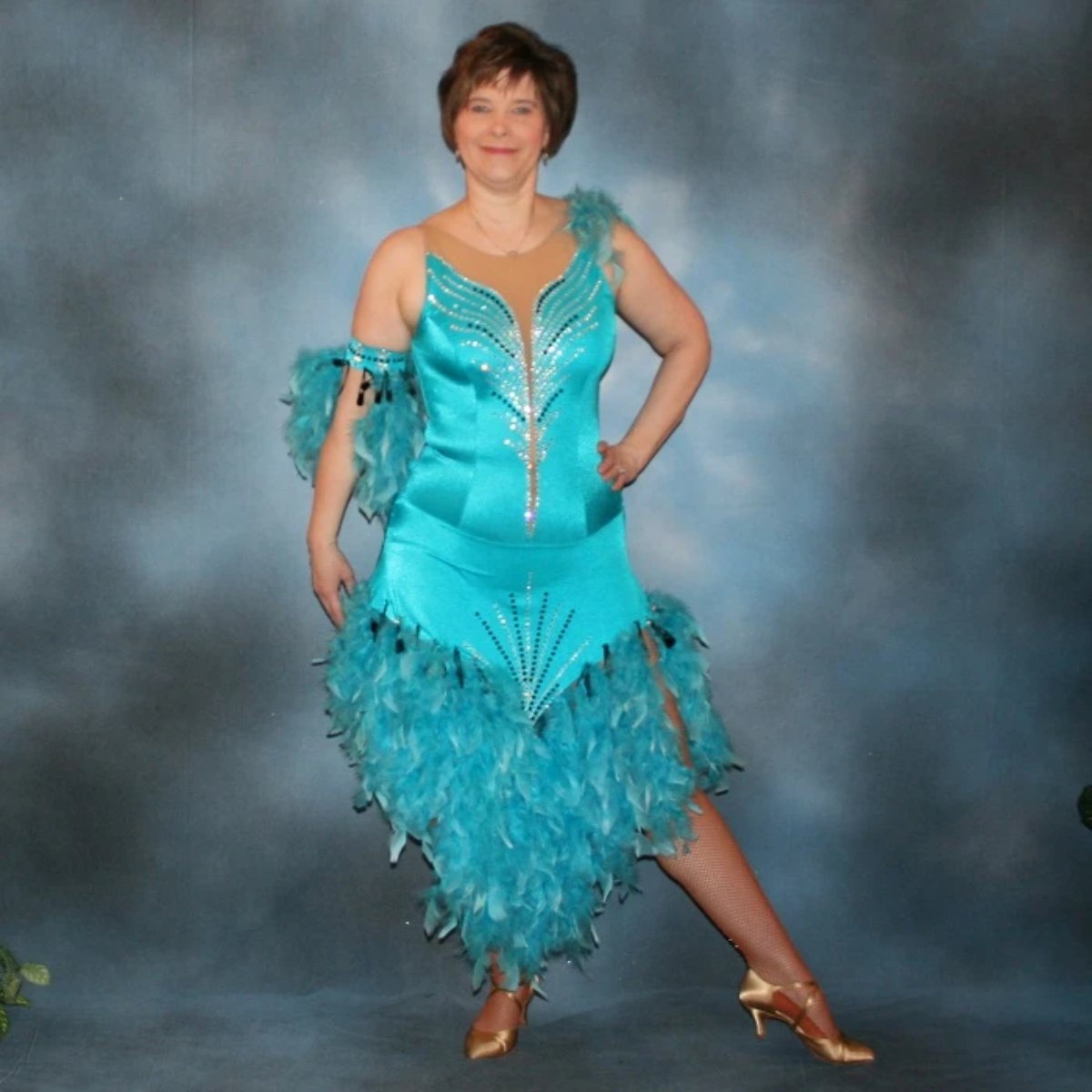 Turquoise Latin/rhythm dance dress created in turquoise lycra on nude illusion, is embellished with crystal and jet black Swarovski rhinestones, with chandelle feathers and black spangles. The v styled skirt slits up high on both sides. Matching arm bands complete the look.