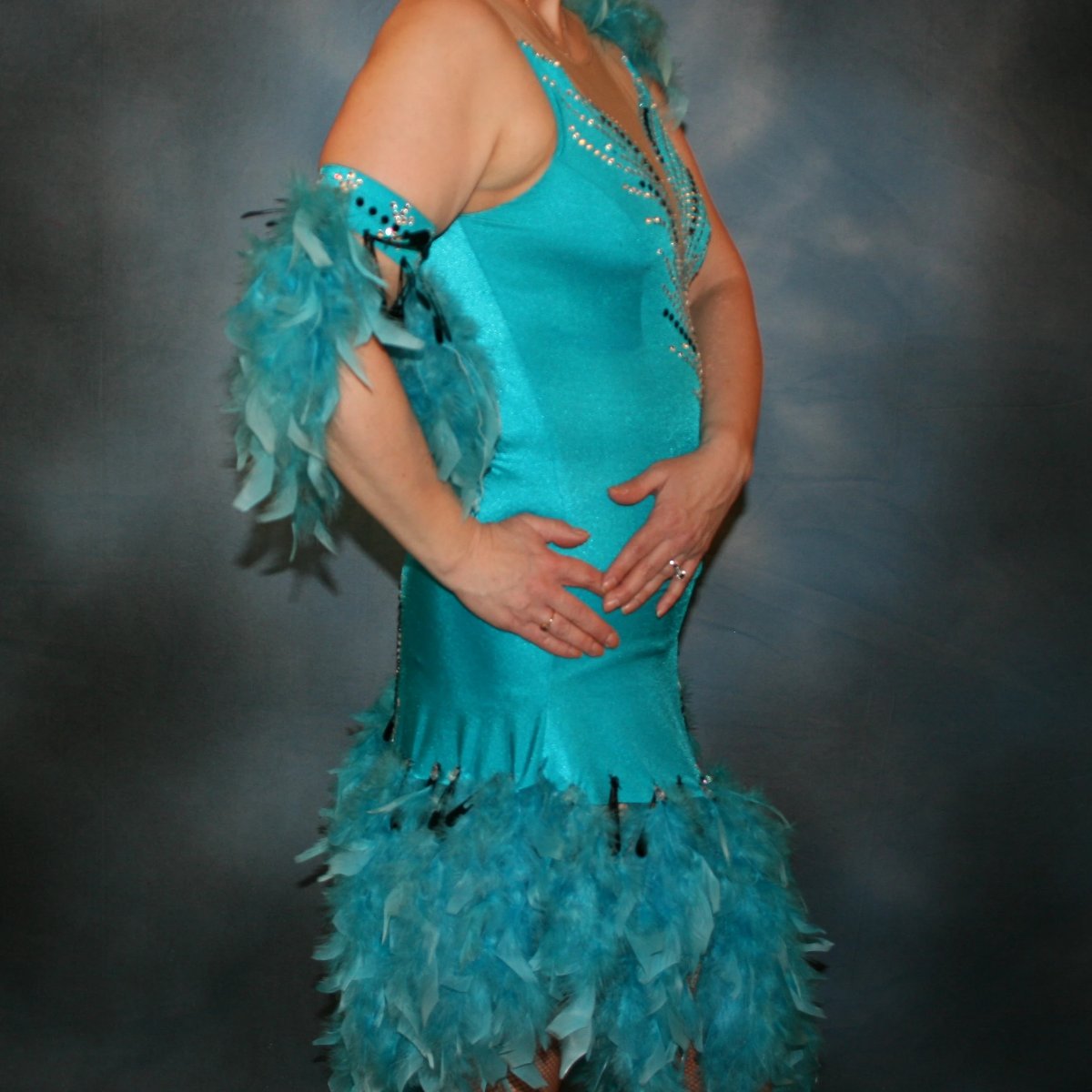 close side view of Turquoise Latin/rhythm dance dress created in turquoise lycra on nude illusion, is embellished with crystal and jet black Swarovski rhinestones, with chandelle feathers and black spangles. The v styled skirt slits up high on both sides. Matching arm bands complete the look.