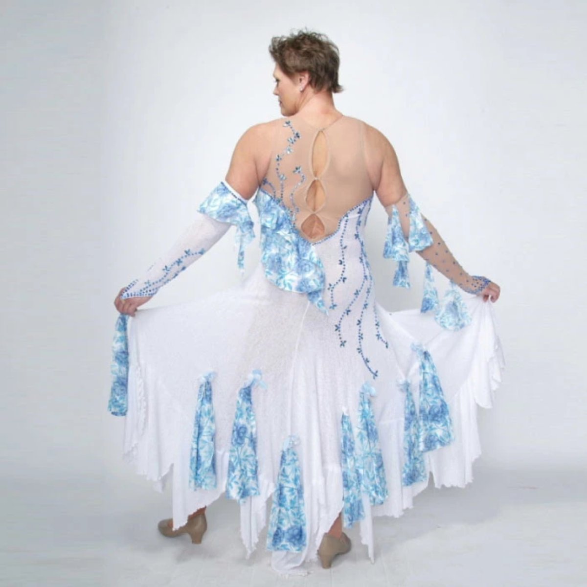 back view of White ballroom dress with blue accents was created on nude illusion of a soft white knit with flounce accents of sky blue & white floral print satin, embellished with sapphire Swarovski stonework & miniature white silk roses. 