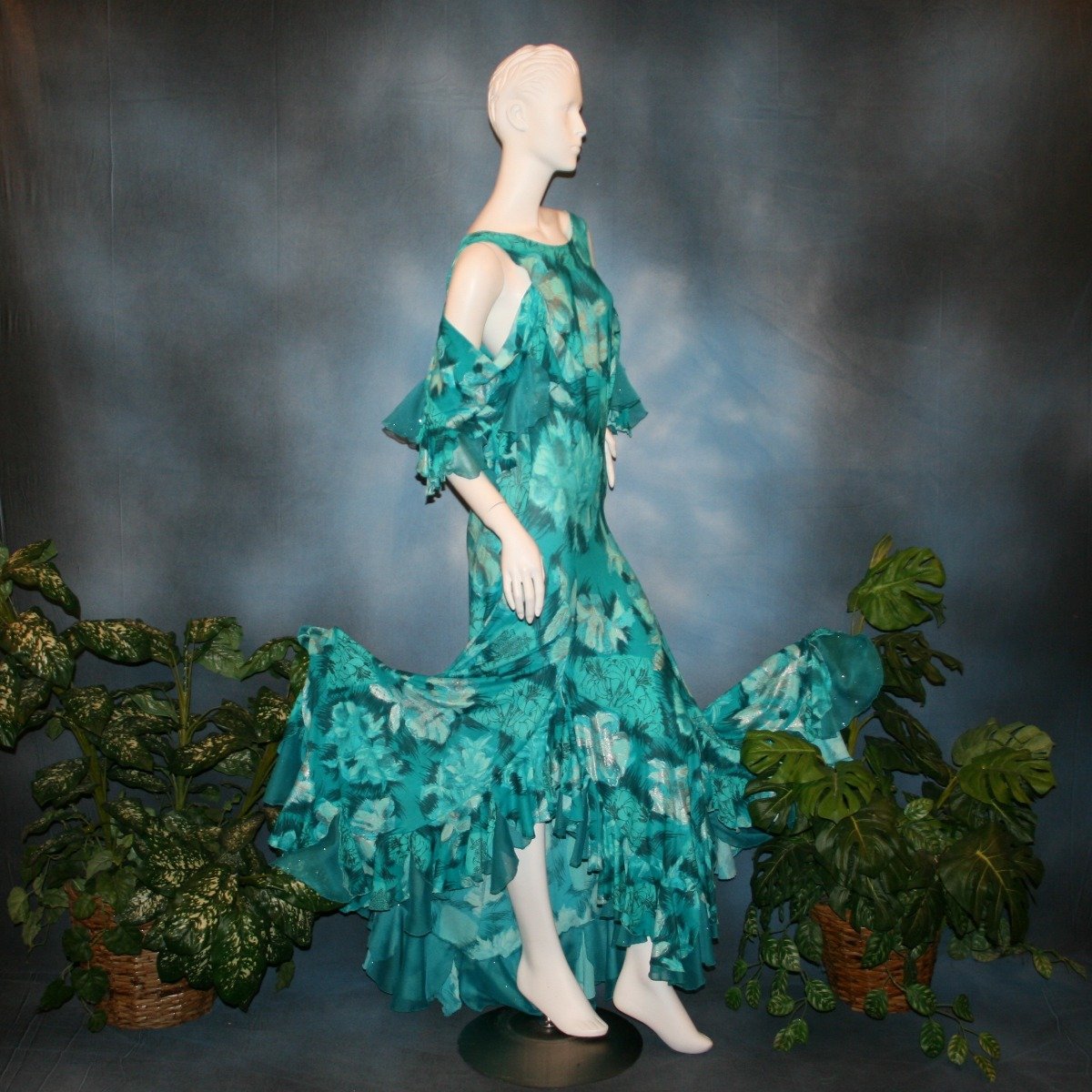 side view of Gorgeous teal tropical print social ballroom dress created in tropical lycra print in shades of teal with a bit of silver sheen accents...absolutely gorgeous fabric! This social ballroom dress features very full skirting with 2 slits & oodles of flounces of the same teal tropical print lycra with intermittent flounces of teal glitter chiffon...along with draping, cold shoulder flounce sleeves...and a low back