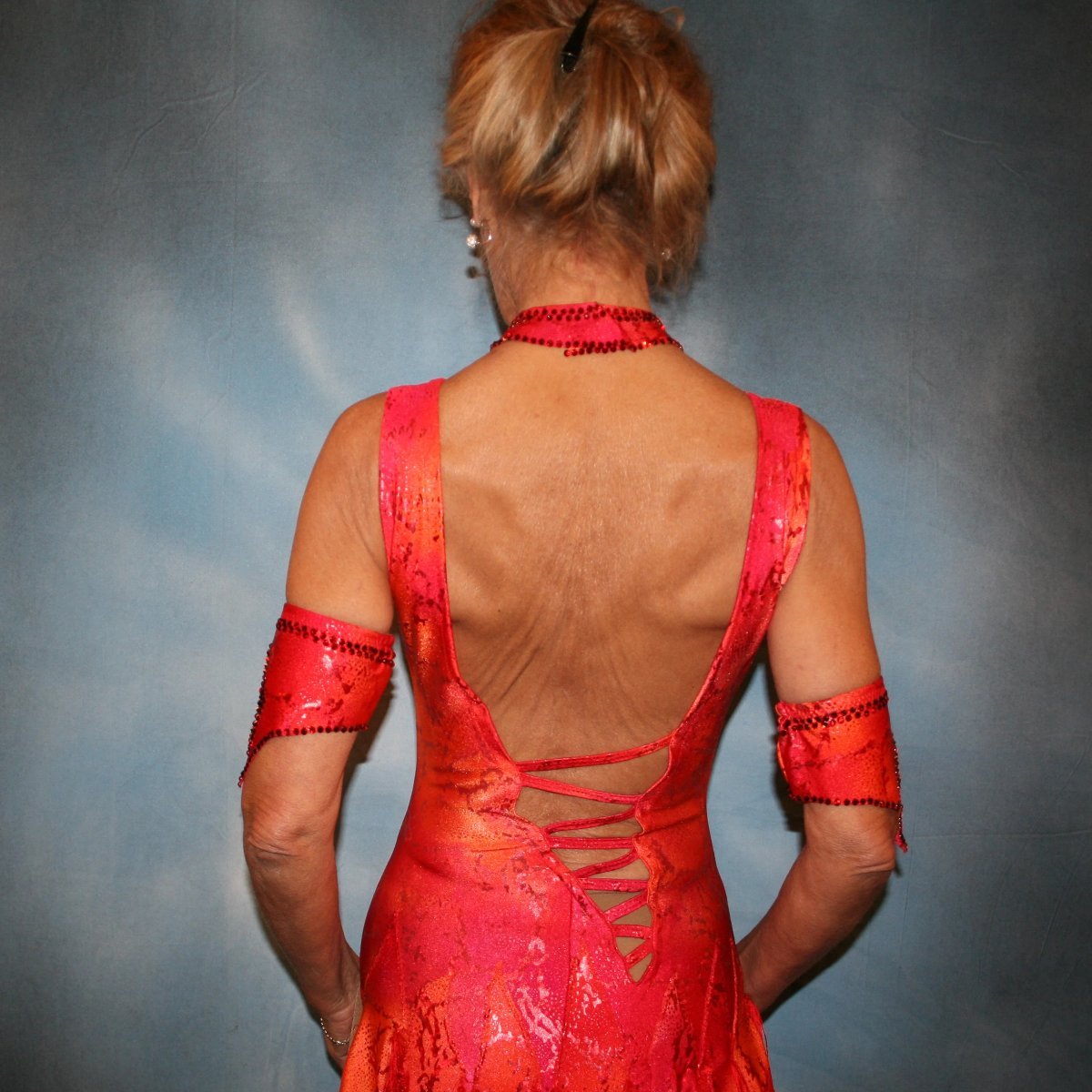 close back view of Red Latin/rhythm dress with fringe, created in a gorgeous Lava hologram metallic lycra of reds, oranges & a touch of yellow, features intricut strapping/lattice detail in design along with several panels bordered with red silky fringe. Neckpiece & arm bands are embellished with siam Swarovski rhinestones.