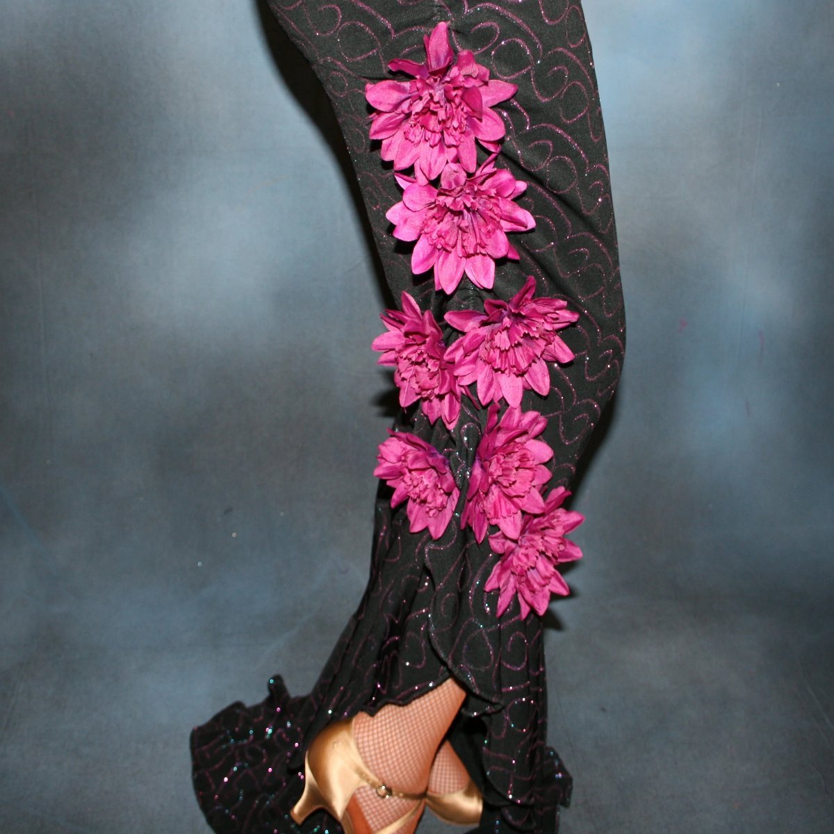 Crystal's Creations close up lower view of Black slinky glitterknit tango dress with glitter scroll design in fuchsia & blue has sleek lines with ruching… embellished with fuschia silk flowers on shoulder & at lower hip & slit in skirt. A fuchsia Swarovski rhinestone is embedded in each flower…stunning on the dance floor!