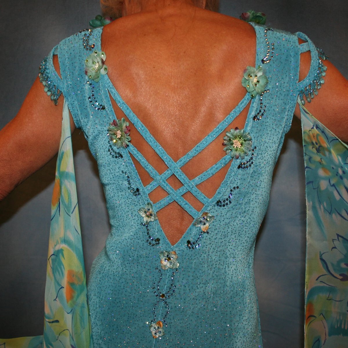 Crystal's Creations close up back view of Turquoise ballroom dress of turquoise glitter slinky features lattice work detailing with yards & yards of print chiffon, enhanced with velveteen flowers plus aquamarine Swarovski rhinestones, hand beading on shoulder detailing... with detachable floats.