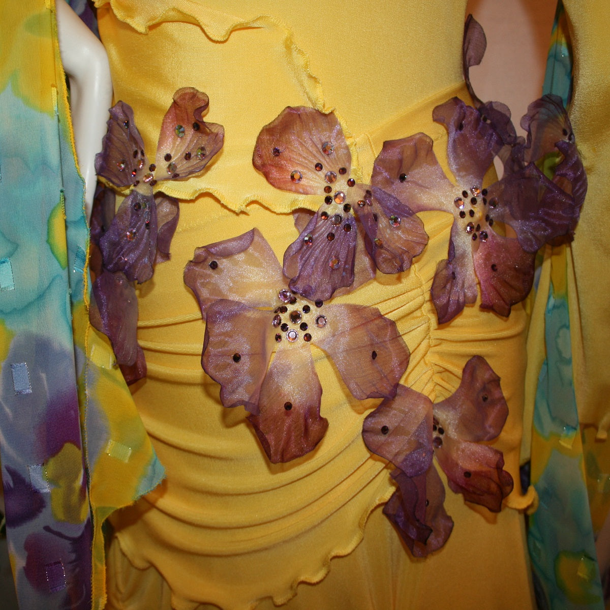 Crystal's Creations clos detail view of Yellow ballroom dress created of luxurious yellow slinky with yards of flower printed textured chiffon of yellow, orchids & blues is embellished with deep orchid silk flowers that have rhinestone work in shades of orchid.