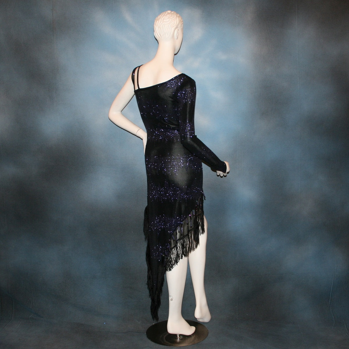 back view of Latin/rhythm/tango dress created in black glitter slinky with an awesome electrifying tanzinite/perwinkle glitter pattern! It features one long sleeve, lattice work detailing up the left side, a short skirt line in front that angles down to a long peak in the back, with a sheer mesh inset, fringe & Swarovski hand beading in tanzanite & black.