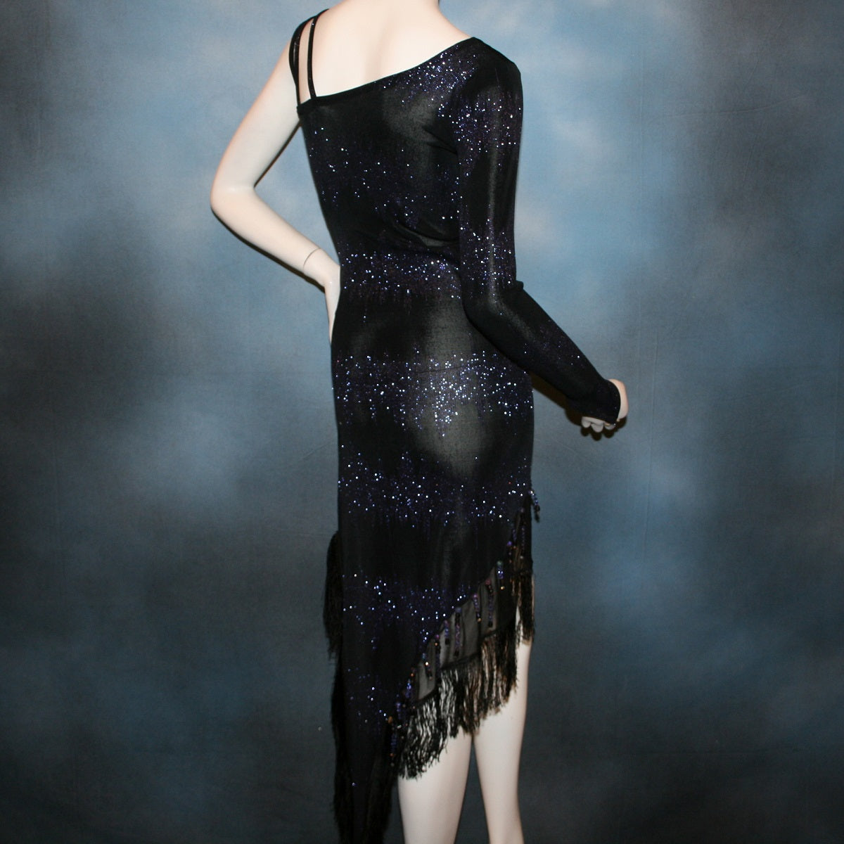 close back view of Latin/rhythm/tango dress created in black glitter slinky with an awesome electrifying tanzinite/perwinkle glitter pattern! It features one long sleeve, lattice work detailing up the left side, a short skirt line in front that angles down to a long peak in the back, with a sheer mesh inset, fringe & Swarovski hand beading in tanzanite & black.