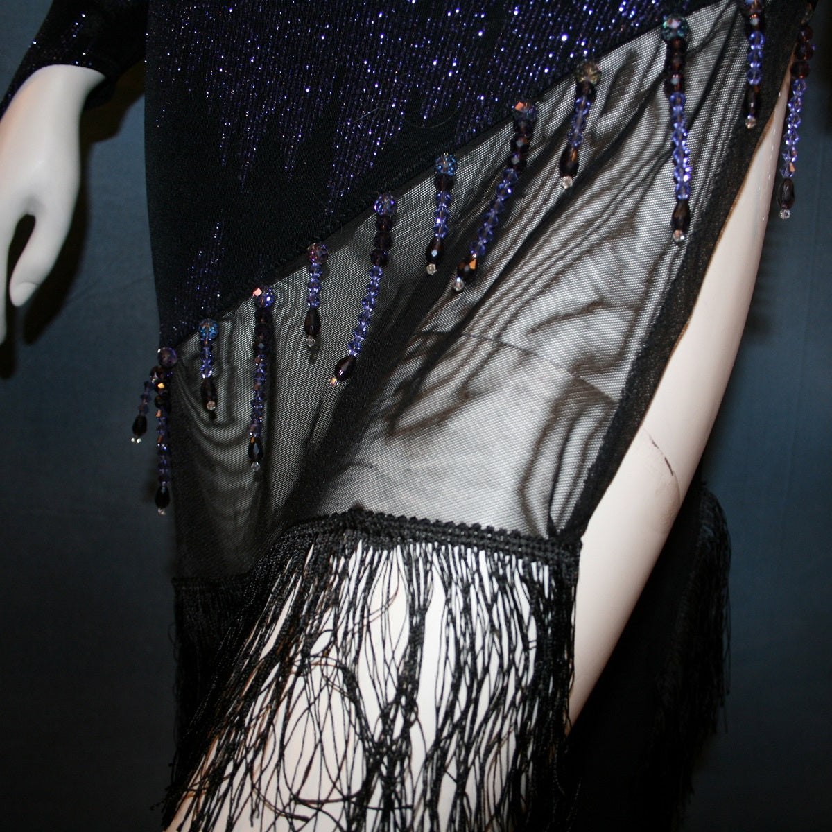 close details of Latin/rhythm/tango dress created in black glitter slinky with an awesome electrifying tanzinite/perwinkle glitter pattern! It features one long sleeve, lattice work detailing up the left side, a short skirt line in front that angles down to a long peak in the back, with a sheer mesh inset, fringe & Swarovski hand beading in tanzanite & black.