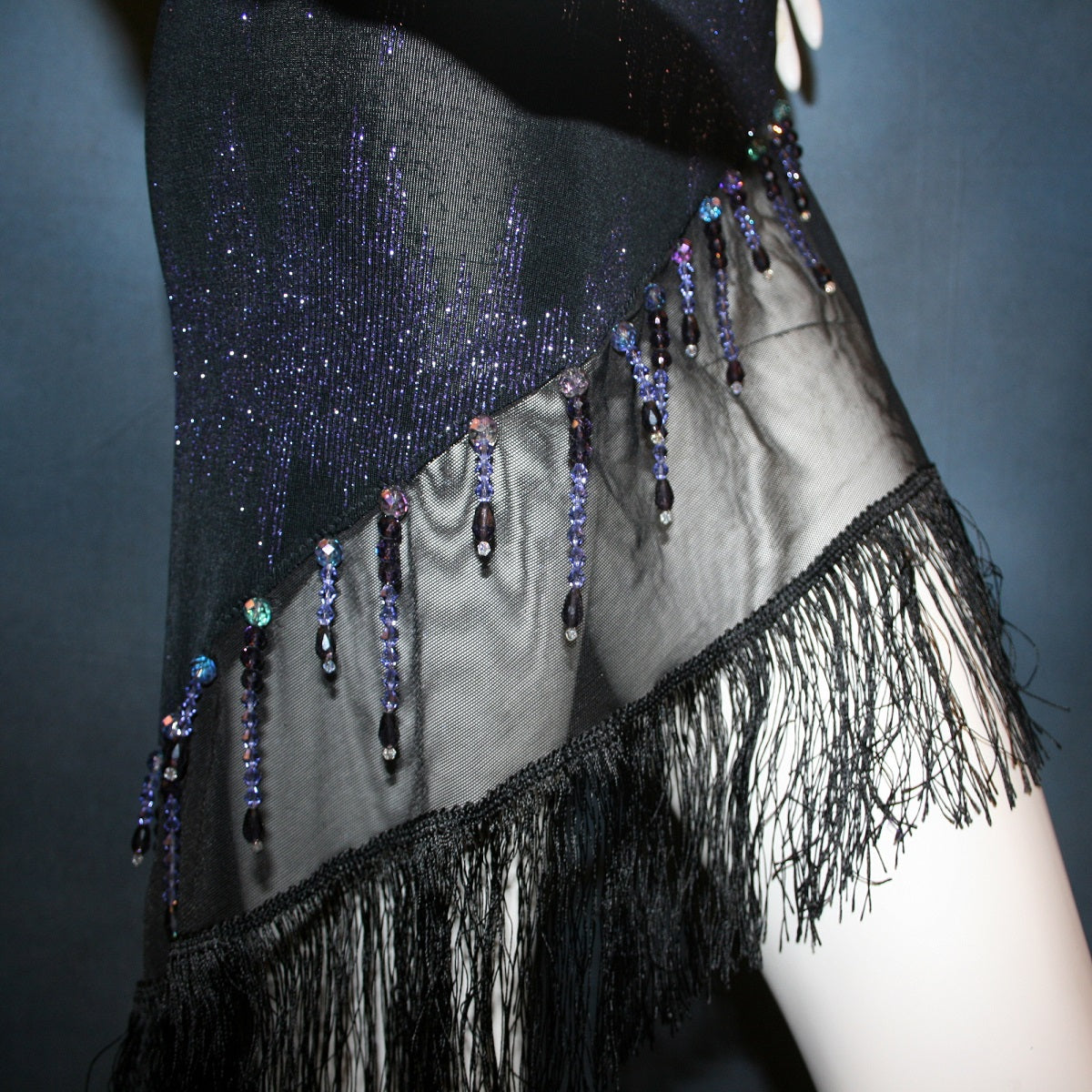 side close details of Latin/rhythm/tango dress created in black glitter slinky with an awesome electrifying tanzinite/perwinkle glitter pattern! It features one long sleeve, lattice work detailing up the left side, a short skirt line in front that angles down to a long peak in the back, with a sheer mesh inset, fringe & Swarovski hand beading in tanzanite & black.