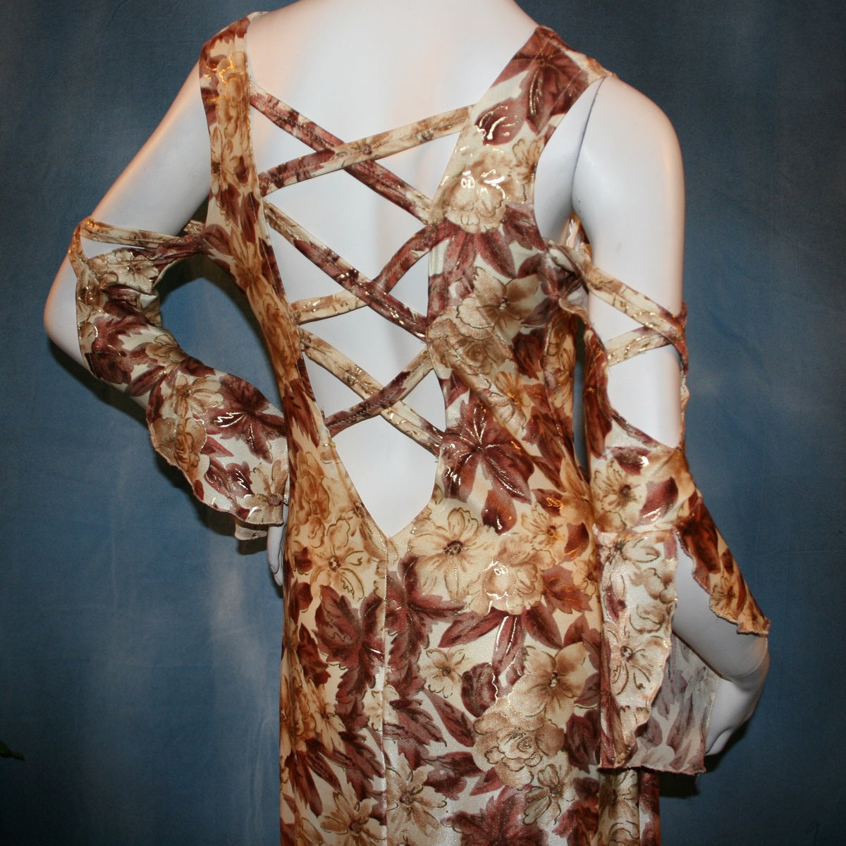 close back view of Ballroom social dress created of brown tropical print lycra with shiny gold accents through the print, features low back & cold shoulder 3/4 sleeves with flounces & lattice strap details, & lots of flounces through the full skirting